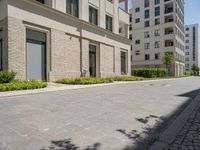 this sidewalk has multiple pavings near two buildings on the sides of it and shrubs beside the curb