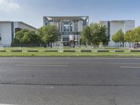 a large gray building sits beside a tree filled road in front of it with traffic going by