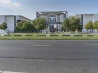 a large gray building sits beside a tree filled road in front of it with traffic going by