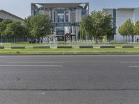 a large gray building sits beside a tree filled road in front of it with traffic going by