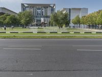 a large gray building sits beside a tree filled road in front of it with traffic going by