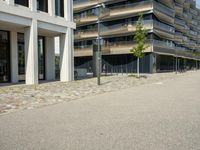 a building with a couple of people riding bikes on the sidewalk below it with cobbles on a paved road