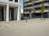 a building with a couple of people riding bikes on the sidewalk below it with cobbles on a paved road