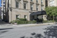 a black awning and the shadow of a tree on the road behind it, across from a tall tan building with windows