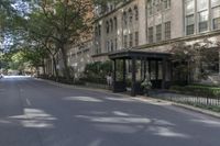 a bench on the side of a street near bushes and buildings'' it has an umbrella over it,