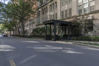 a bench on the side of a street near bushes and buildings'' it has an umbrella over it,