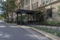a bench on the side of a street near bushes and buildings'' it has an umbrella over it,