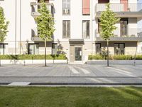 an empty sidewalk in front of a multi - story building near green trees and grass