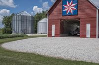 a barn on top of a gravel road next to grass and trees in front of a blue sky