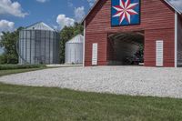 a barn on top of a gravel road next to grass and trees in front of a blue sky