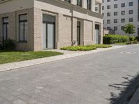 a block paved area with bushes in the front and sidewalk with brick paving and building behind