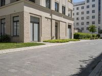 a block paved area with bushes in the front and sidewalk with brick paving and building behind