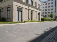 a block paved area with bushes in the front and sidewalk with brick paving and building behind