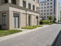 a block paved area with bushes in the front and sidewalk with brick paving and building behind