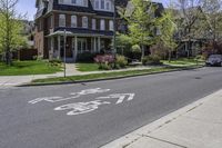 an empty street in front of a large house with bikes written on the sidewalk, in front of houses with trees