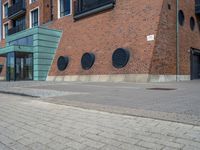 a woman wearing red is walking across the pavement outside her apartment block, which features large circular windows and a brick wall