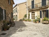 an old stone house with two lawn benches and pots of plants are set in a cobbled sidewalk between two buildings