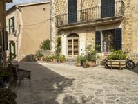 an old stone house with two lawn benches and pots of plants are set in a cobbled sidewalk between two buildings