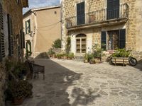 an old stone house with two lawn benches and pots of plants are set in a cobbled sidewalk between two buildings