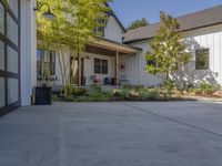 an empty driveway and a large white house with windows and patio, surrounded by large trees and shrubs