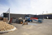 a big blue crane in a lot with some other pallets around it and the building is grey