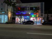 a night scene of two black cars parked in front of a colorful storefront at the corner of a street