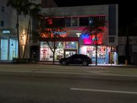 a night scene of two black cars parked in front of a colorful storefront at the corner of a street