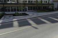 the shadows of a person riding a bike past a building on the street while there is an empty sidewalk in front of them