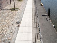 a river side walkway leads to a large boat yard with a few benches and people walking along