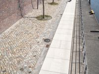 a river side walkway leads to a large boat yard with a few benches and people walking along