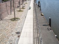 a river side walkway leads to a large boat yard with a few benches and people walking along