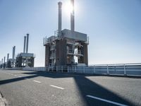 two people walking along a road near large power plants, with water visible from them