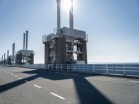 two people walking along a road near large power plants, with water visible from them