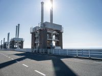 two people walking along a road near large power plants, with water visible from them