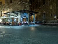 a long street filled with people sitting at tables near tall buildings at night and parked motorcycles