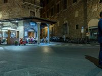 a long street filled with people sitting at tables near tall buildings at night and parked motorcycles