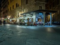 a long street filled with people sitting at tables near tall buildings at night and parked motorcycles