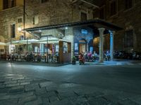 a long street filled with people sitting at tables near tall buildings at night and parked motorcycles