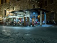 a long street filled with people sitting at tables near tall buildings at night and parked motorcycles