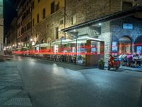 a long street filled with people sitting at tables near tall buildings at night and parked motorcycles