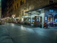 a long street filled with people sitting at tables near tall buildings at night and parked motorcycles