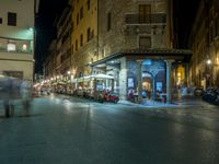 a long street filled with people sitting at tables near tall buildings at night and parked motorcycles