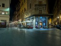 a long street filled with people sitting at tables near tall buildings at night and parked motorcycles