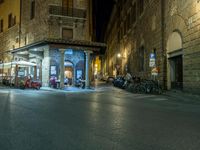 a long street filled with people sitting at tables near tall buildings at night and parked motorcycles