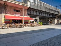 tables and chairs on sidewalk outside restaurant under walkway with traffic light in area above walkway