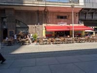 tables and chairs on sidewalk outside restaurant under walkway with traffic light in area above walkway