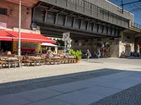 tables and chairs on sidewalk outside restaurant under walkway with traffic light in area above walkway