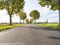 an empty asphalt road lined by trees in the countryside with a church tower to the right of it