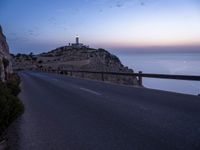 a narrow highway with a lighthouse on a rock cliff above the ocean at dusk overlooking an island