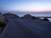 a narrow highway with a lighthouse on a rock cliff above the ocean at dusk overlooking an island